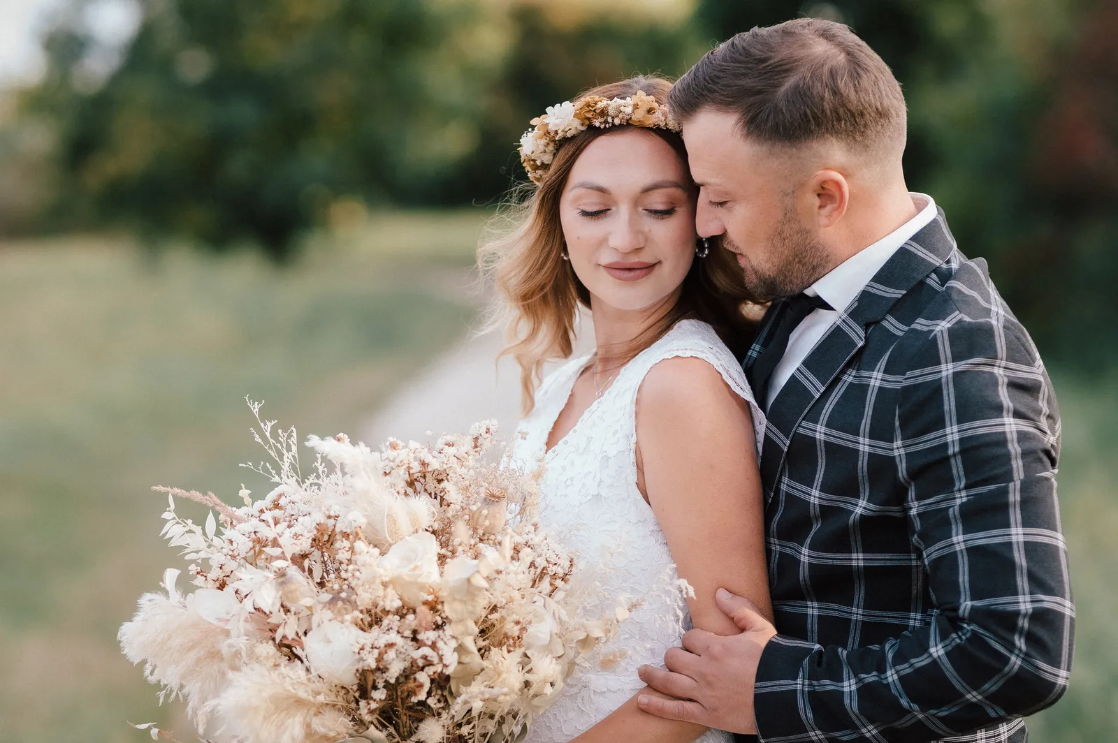 Braut mit Blumenkranz und Trockenblumenstrauß, der Bräutigam küsst ihre Schläfe
